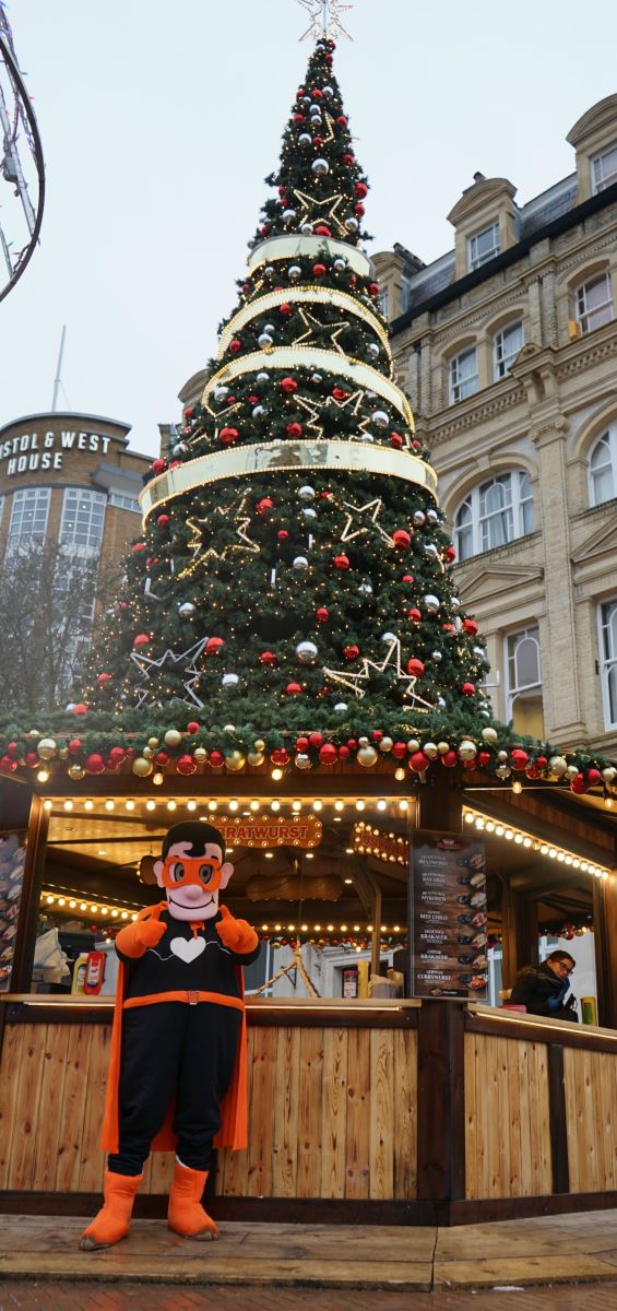 Mr Super Health-on-Line standing in front of the Bratwurst stand at the Bournemouth Christmas Tree Wonderland market, with two thumbs up in his black suit and orange cape